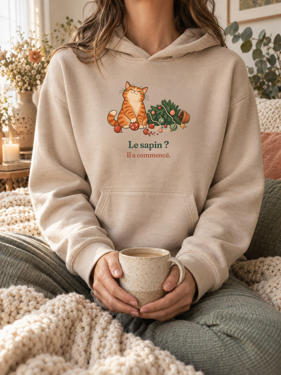 Visuel d’illustration – sweat à capuche sable porté, une tasse entre les mains, dans un décor cocooning, avec le motif de Noël : un chat roux assis près d’un sapin de Noël renversé et la phrase « Le sapin ? Il a commencé. »
