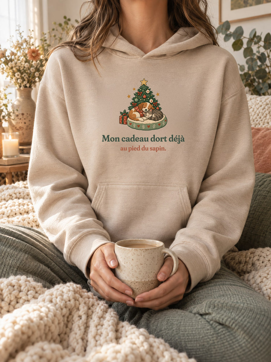 Visuel d’illustration – sweat à capuche sable porté, une tasse entre les mains, dans un décor cocooning, avec le motif de Noël : un chien et un chat endormis au pied d’un petit sapin et la phrase « Mon cadeau dort déjà au pied du sapin. »