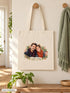 Visuel d’illustration – tote bag accroché à une patère en bois dans une entrée, écharpe et plante verte, avec un portrait de couple fictif en style Portrait signature, prénoms « Camille & Hugo »