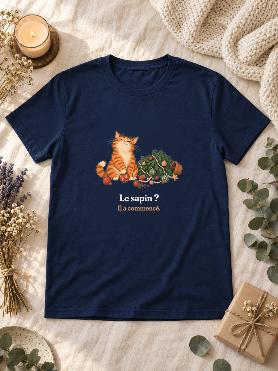 Visuel d’illustration – t-shirt navy posé à plat sur du lin, avec des fleurs séchées, une bougie et un paquet cadeau, avec le motif de Noël : un chat roux assis près d’un sapin de Noël renversé et la phrase « Le sapin ? Il a commencé. »