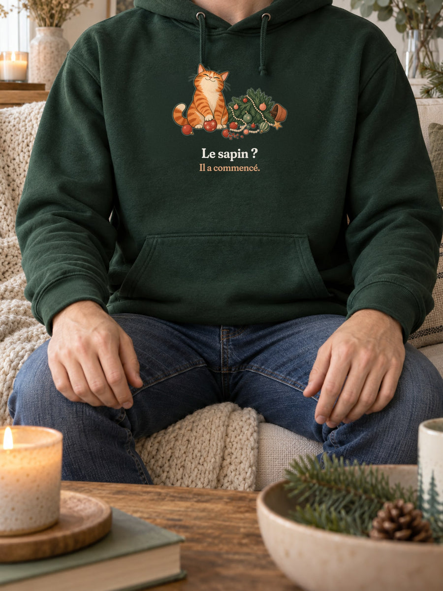 Visuel d’illustration – sweat à capuche vert forêt porté, sur un canapé, près d’une bougie, avec le motif de Noël : un chat roux assis près d’un sapin de Noël renversé et la phrase « Le sapin ? Il a commencé. »