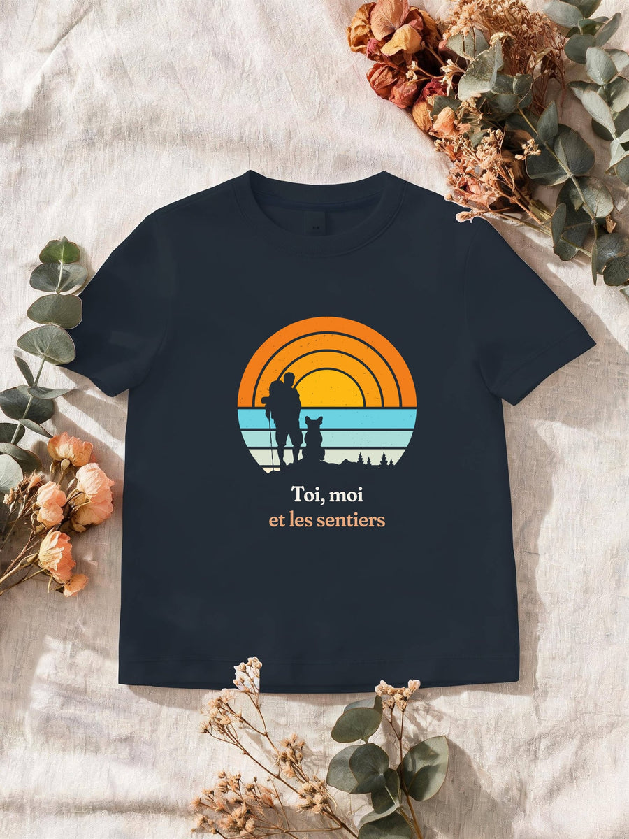 Visuel d’illustration – T-shirt homme navy avec le motif un randonneur et son chien assis, en silhouettes devant un grand soleil orange à bandes rétro et « Toi, moi et les sentiers »