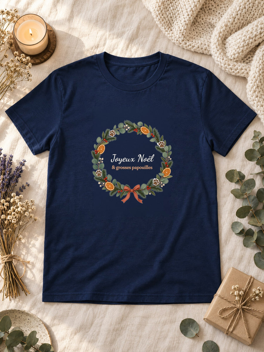 Visuel d’illustration – t-shirt navy posé à plat sur du lin, avec des fleurs séchées, une bougie et un paquet cadeau, avec le motif de Noël : une couronne de Noël avec des empreintes de pattes et le texte « Joyeux Noël & grosses papouilles »