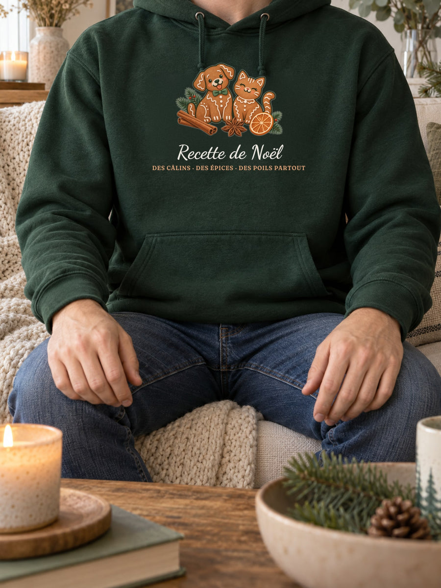 Visuel d’illustration – sweat à capuche vert forêt porté, sur un canapé, près d’une bougie, avec le motif de Noël : deux biscuits de pain d’épices en forme de chien et de chat et le texte « Recette de Noël : des câlins, des épices, des poils partout »