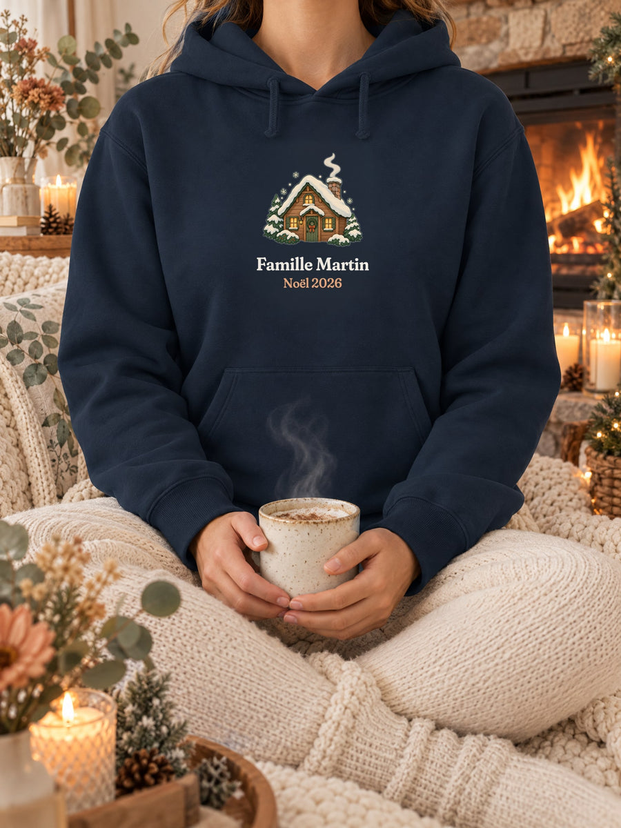 Visuel d’illustration – sweat à capuche navy dans une mise en situation, avec le motif un chalet sous la neige et « Famille Martin · Noël 2026 » (exemple de nom)