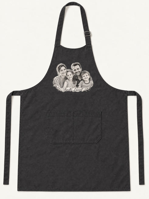 Visuel d’illustration – tablier de cuisine noir en coton, à plat, vue de face, avec un portrait de famille fictive au crayon clair, style Croquis, texte « Famille Laurent »