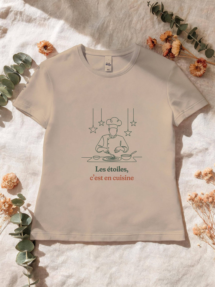 Visuel d’illustration – T-shirt femme sable avec le motif un cuisinier en toque qui dresse une assiette sous des étoiles suspendues, dessiné d’un seul trait et « Les étoiles, c’est en cuisine »