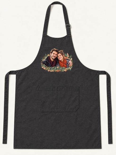 Visuel d’illustration – tablier de cuisine noir en coton, à plat, vue de face, avec un portrait de couple fictif en style Portrait signature, prénoms clairs « Camille & Hugo »