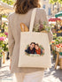 Visuel d’illustration – tote bag porté à l’épaule au marché, vu de dos, avec une baguette et des fleurs, avec un portrait de couple fictif en style Portrait signature, prénoms « Camille & Hugo »