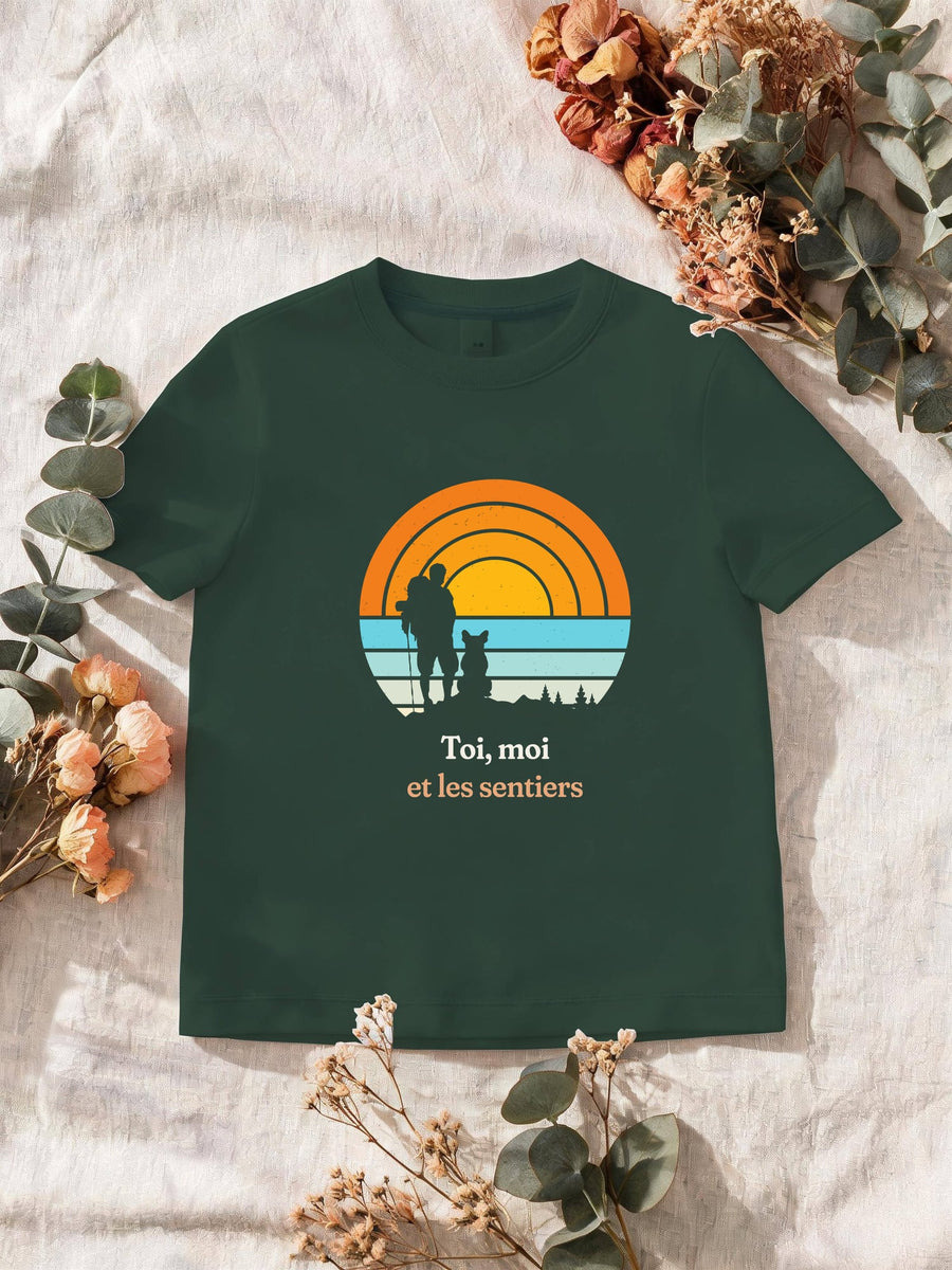 Visuel d’illustration – T-shirt homme vert forêt avec le motif un randonneur et son chien assis, en silhouettes devant un grand soleil orange à bandes rétro et « Toi, moi et les sentiers »