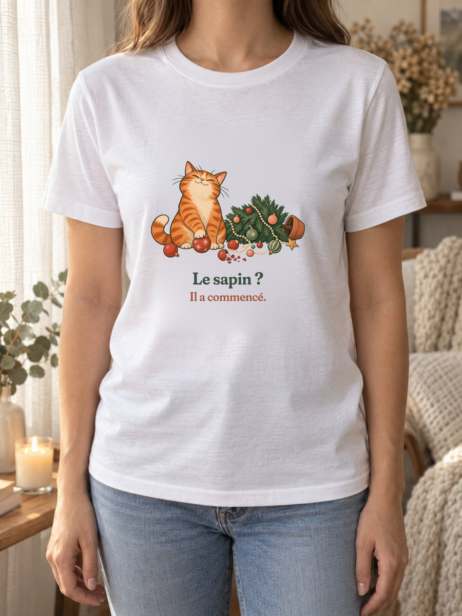 Visuel d’illustration – t-shirt blanc porté, près d’un rideau de lin et d’une bougie, avec le motif de Noël : un chat roux assis près d’un sapin de Noël renversé et la phrase « Le sapin ? Il a commencé. »