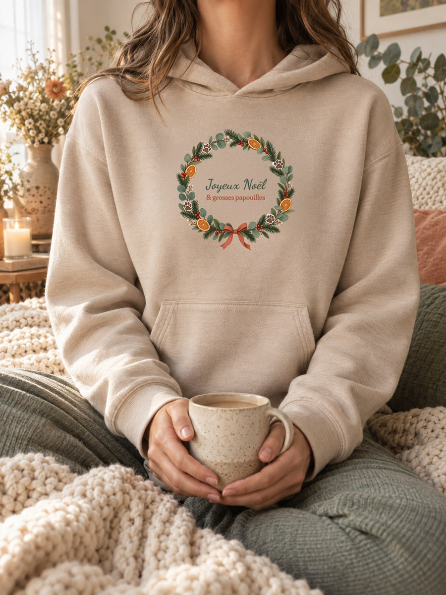Visuel d’illustration – sweat à capuche sable porté, une tasse entre les mains, dans un décor cocooning, avec le motif de Noël : une couronne de Noël avec des empreintes de pattes et le texte « Joyeux Noël & grosses papouilles »