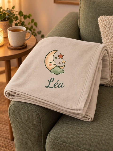 Visuel d’illustration – plaid plié sur l’accoudoir d’un canapé vert sauge, imprimé d’une lune endormie et « Léa » (exemple)