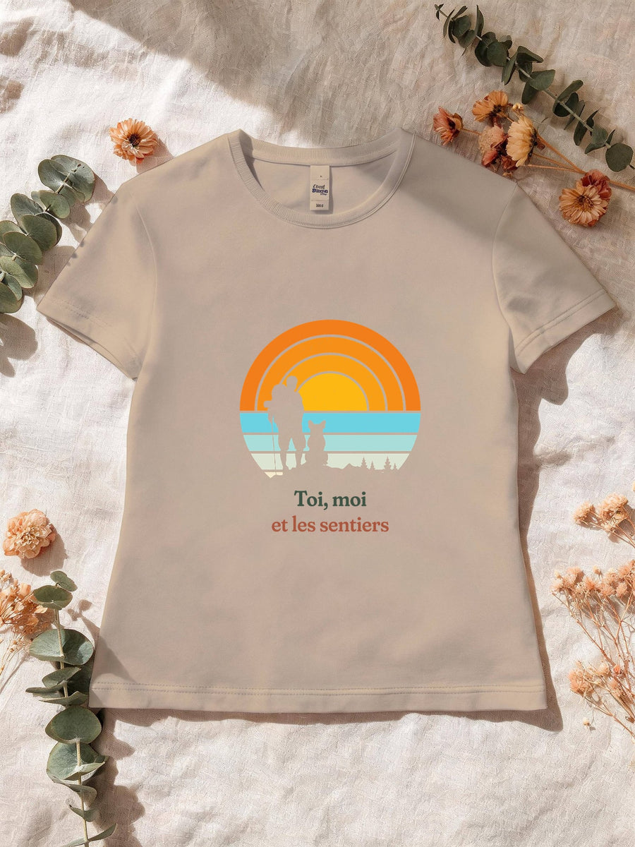 Visuel d’illustration – T-shirt femme sable avec le motif un randonneur et son chien assis, en silhouettes devant un grand soleil orange à bandes rétro et « Toi, moi et les sentiers »