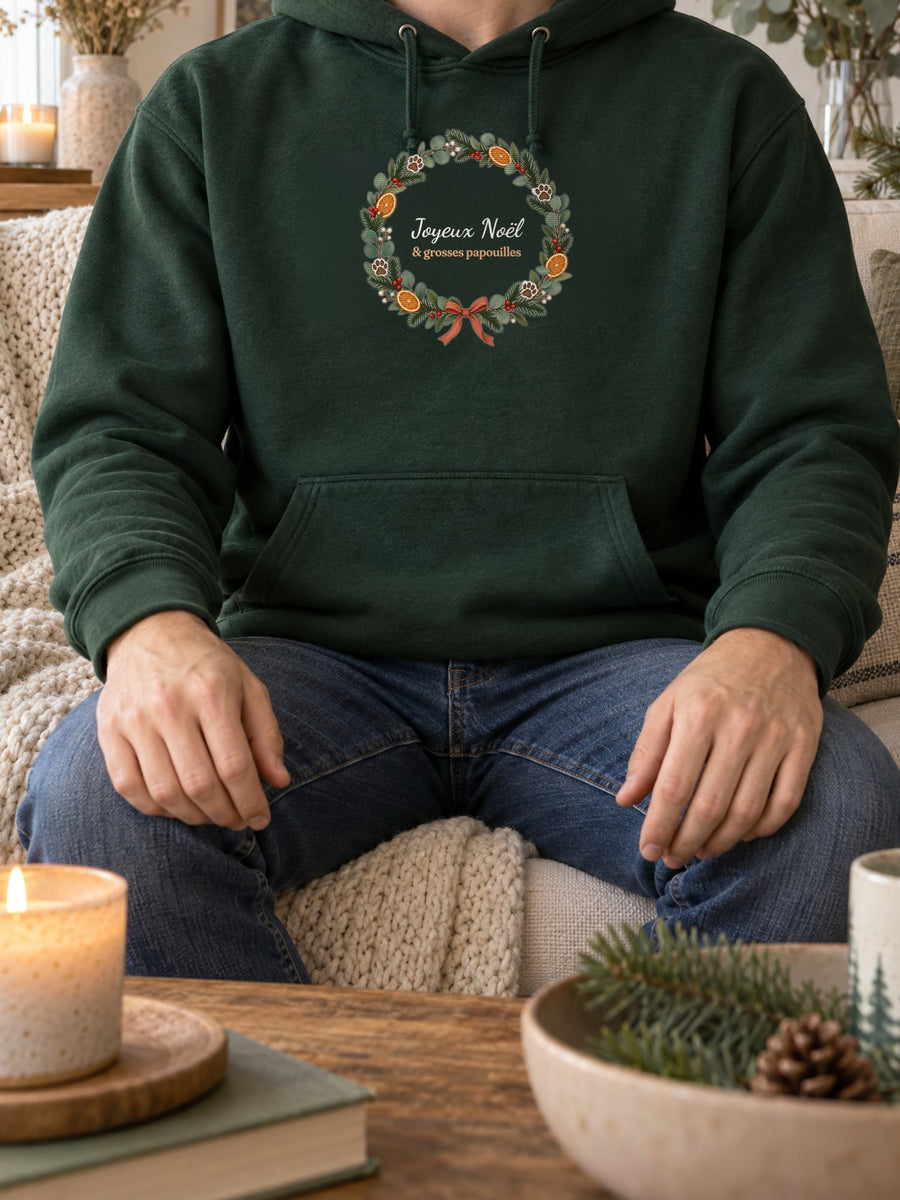 Visuel d’illustration – sweat à capuche vert forêt porté, sur un canapé, près d’une bougie, avec le motif de Noël : une couronne de Noël avec des empreintes de pattes et le texte « Joyeux Noël & grosses papouilles »