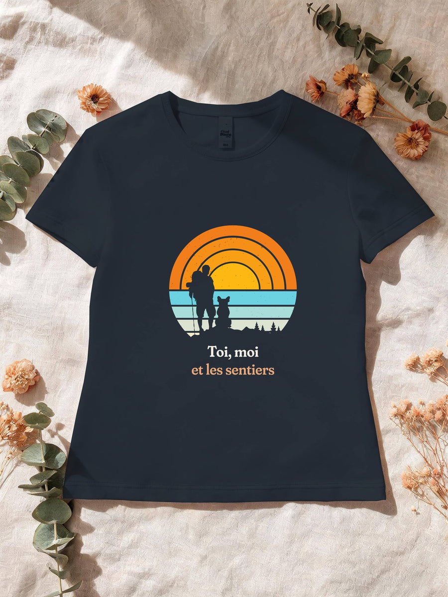 Visuel d’illustration – T-shirt femme navy avec le motif un randonneur et son chien assis, en silhouettes devant un grand soleil orange à bandes rétro et « Toi, moi et les sentiers »