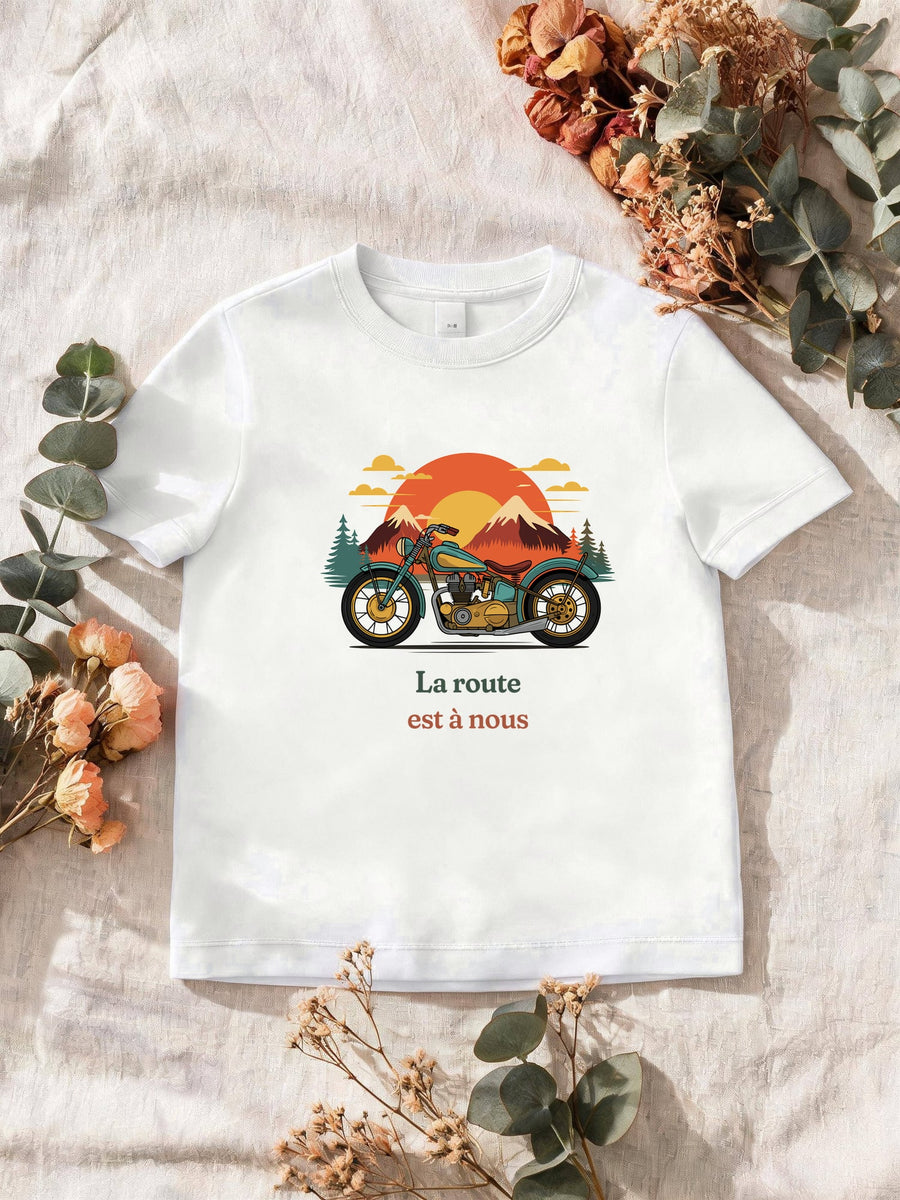 Visuel d’illustration – T-shirt homme blanc avec le motif une moto ancienne illustrée en couleurs devant des montagnes, des sapins et un soleil couchant et « La route est à nous »
