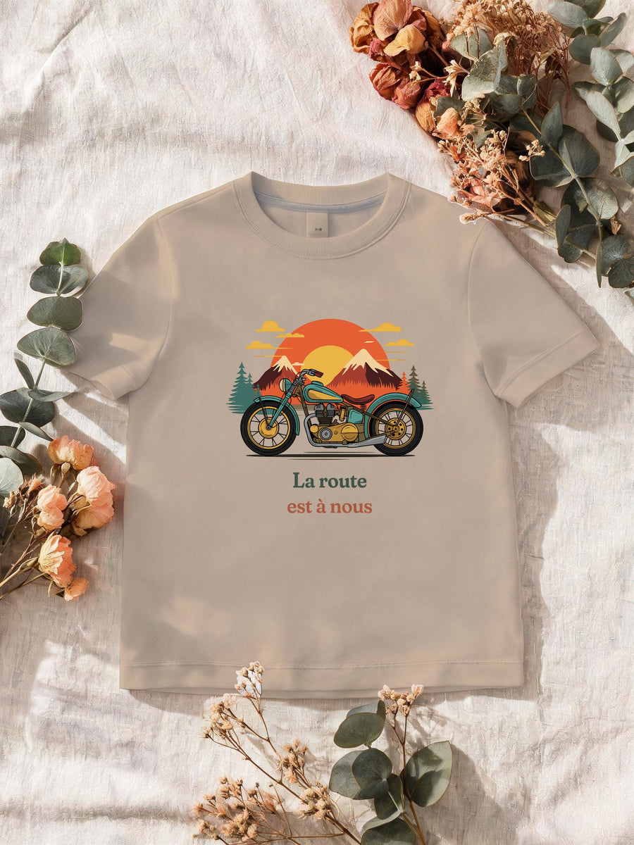 Visuel d’illustration – T-shirt homme sable avec le motif une moto ancienne illustrée en couleurs devant des montagnes, des sapins et un soleil couchant et « La route est à nous »