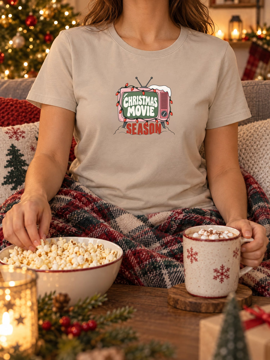 Visuel d’illustration – mise en situation : un t-shirt femme sable, sous un plaid avec du pop-corn, avec la télé rétro rose et sa guirlande, « Christmas Movie Season »