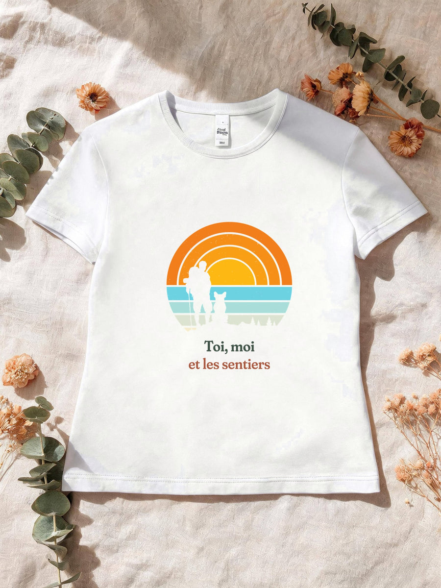 Visuel d’illustration – T-shirt femme blanc avec le motif un randonneur et son chien assis, en silhouettes devant un grand soleil orange à bandes rétro et « Toi, moi et les sentiers »