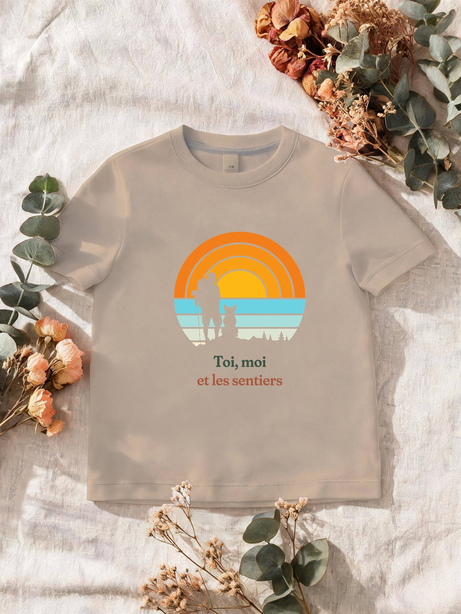 Visuel d’illustration – T-shirt homme sable avec le motif un randonneur et son chien assis, en silhouettes devant un grand soleil orange à bandes rétro et « Toi, moi et les sentiers »