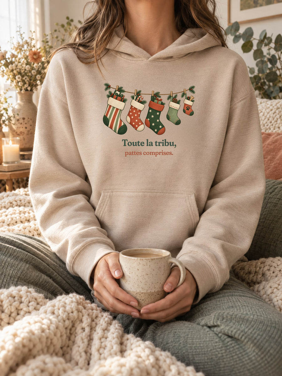 Visuel d’illustration – sweat à capuche sable porté, une tasse entre les mains, dans un décor cocooning, avec le motif de Noël : une guirlande de chaussettes de Noël, dont une petite avec une empreinte de patte, et la phrase « Toute la tribu, pattes comprises. »