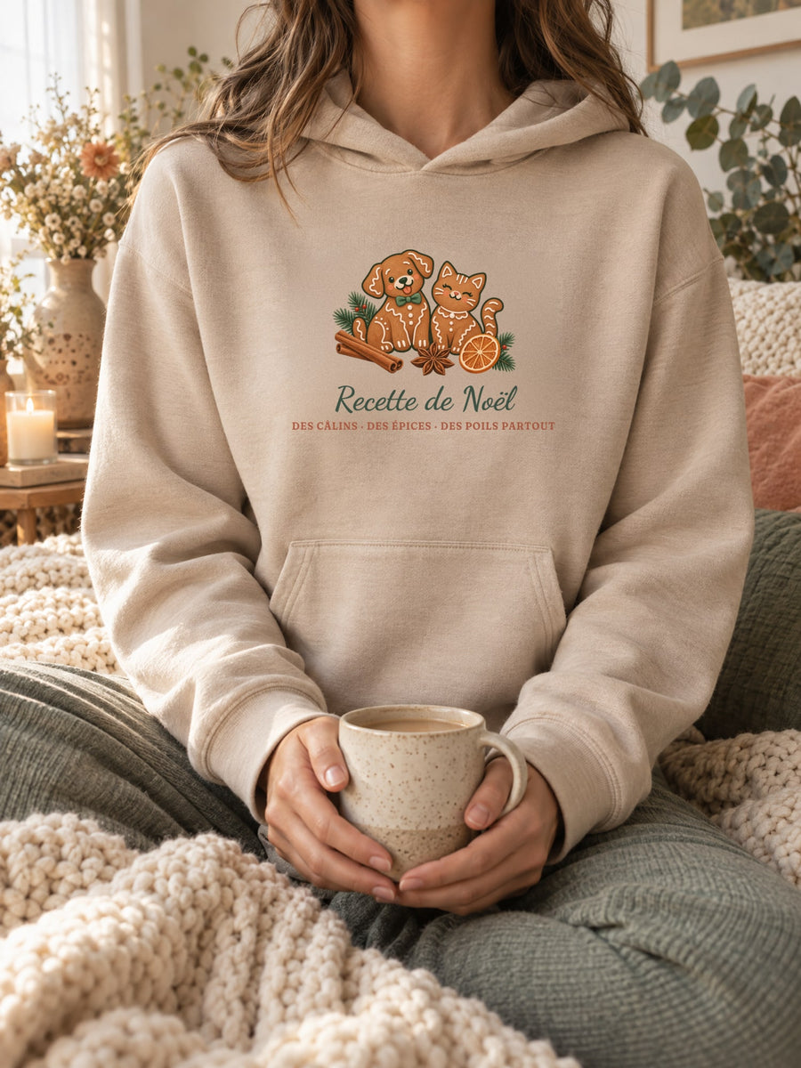 Visuel d’illustration – sweat à capuche sable porté, une tasse entre les mains, dans un décor cocooning, avec le motif de Noël : deux biscuits de pain d’épices en forme de chien et de chat et le texte « Recette de Noël : des câlins, des épices, des poils partout »