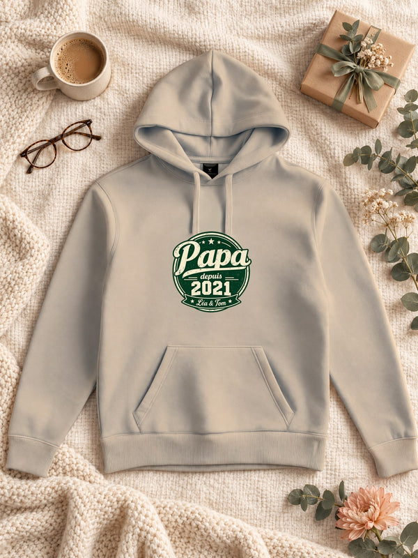 Visuel d’illustration – Sweat à capuche sable avec le design « Papa depuis… », posé à plat sur un plaid, avec les prénoms d’exemple  et l’année 2021