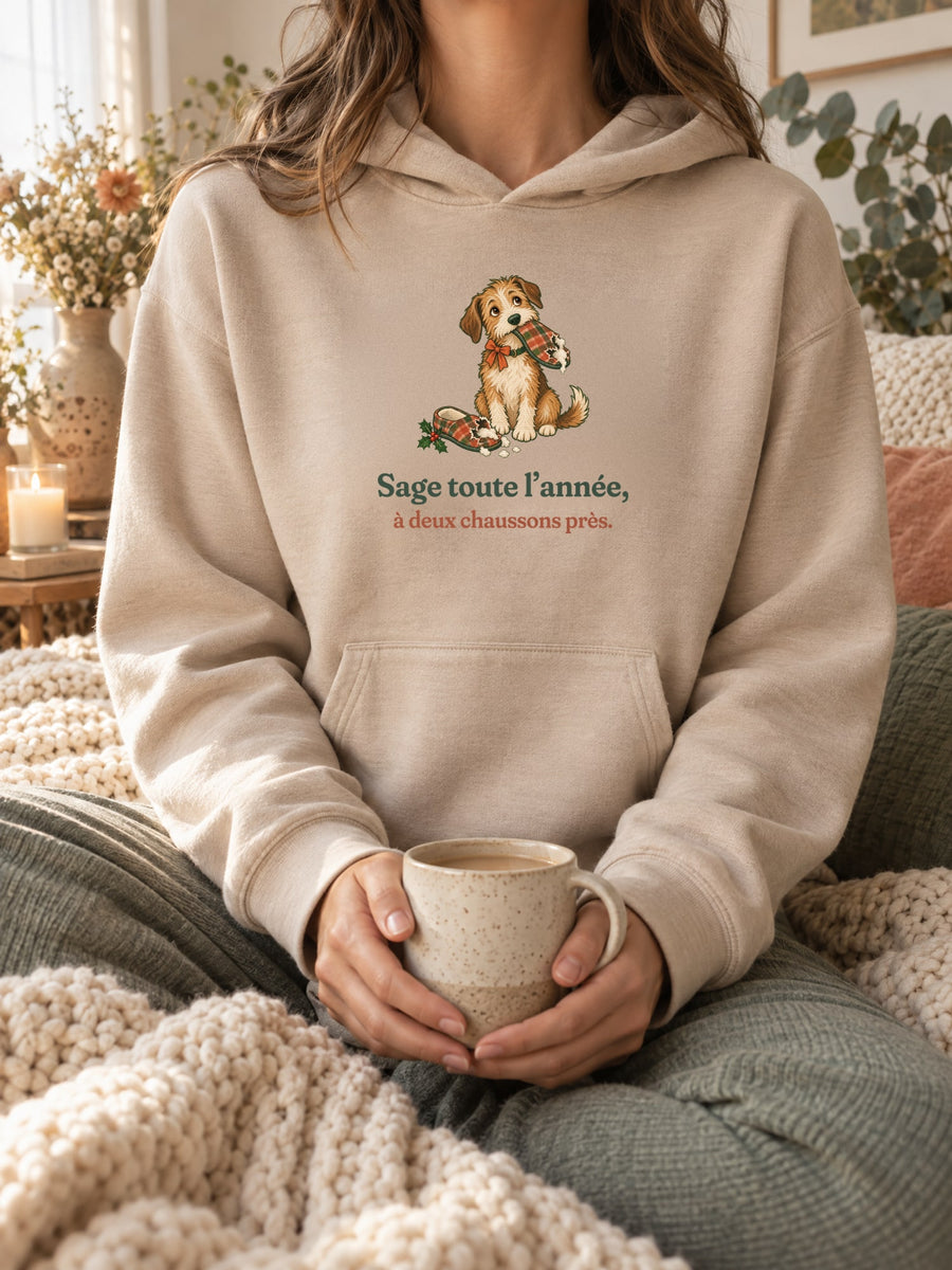 Visuel d’illustration – sweat à capuche sable porté, une tasse entre les mains, dans un décor cocooning, avec le motif de Noël : un chien assis, un chausson dans la gueule, et la phrase « Sage toute l’année, à deux chaussons près. »