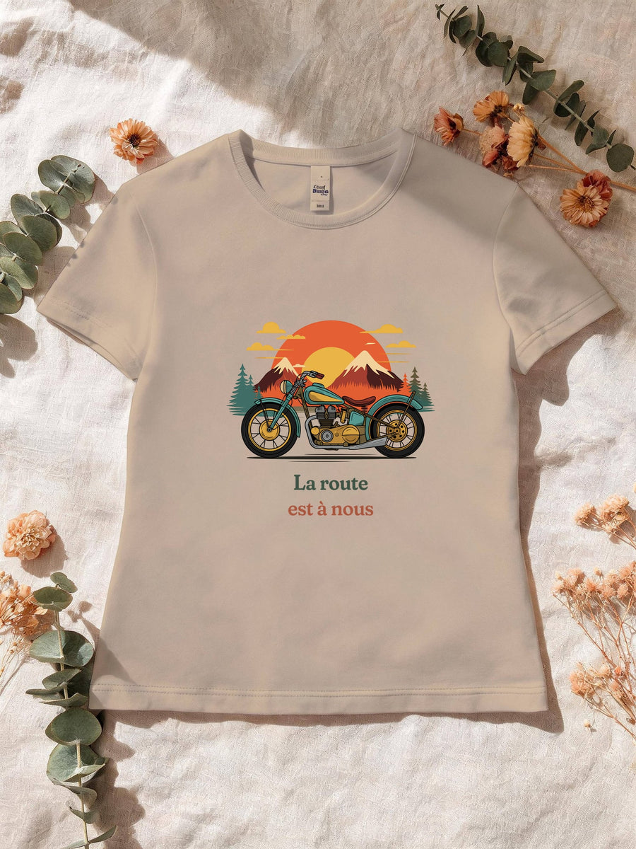 Visuel d’illustration – T-shirt femme sable avec le motif une moto ancienne illustrée en couleurs devant des montagnes, des sapins et un soleil couchant et « La route est à nous »