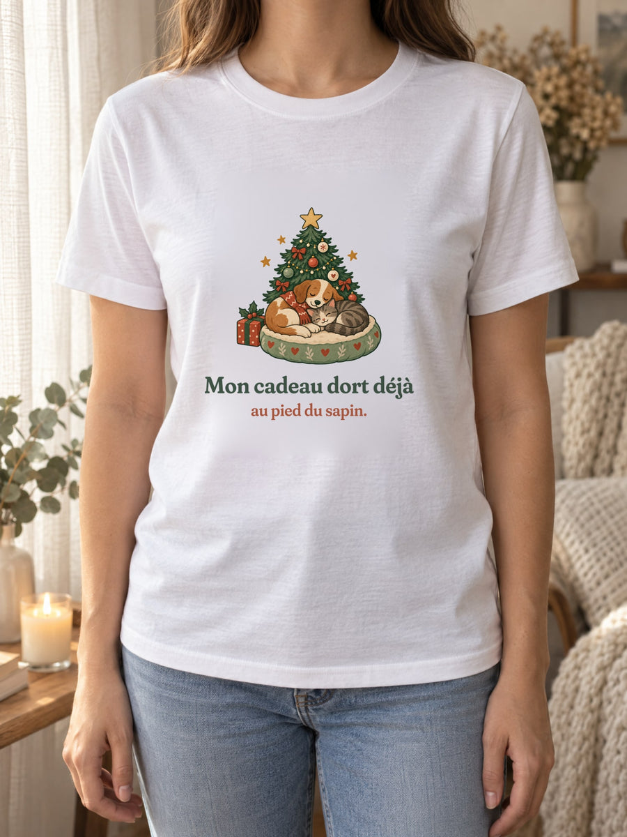 Visuel d’illustration – t-shirt blanc porté, près d’un rideau de lin et d’une bougie, avec le motif de Noël : un chien et un chat endormis au pied d’un petit sapin et la phrase « Mon cadeau dort déjà au pied du sapin. »