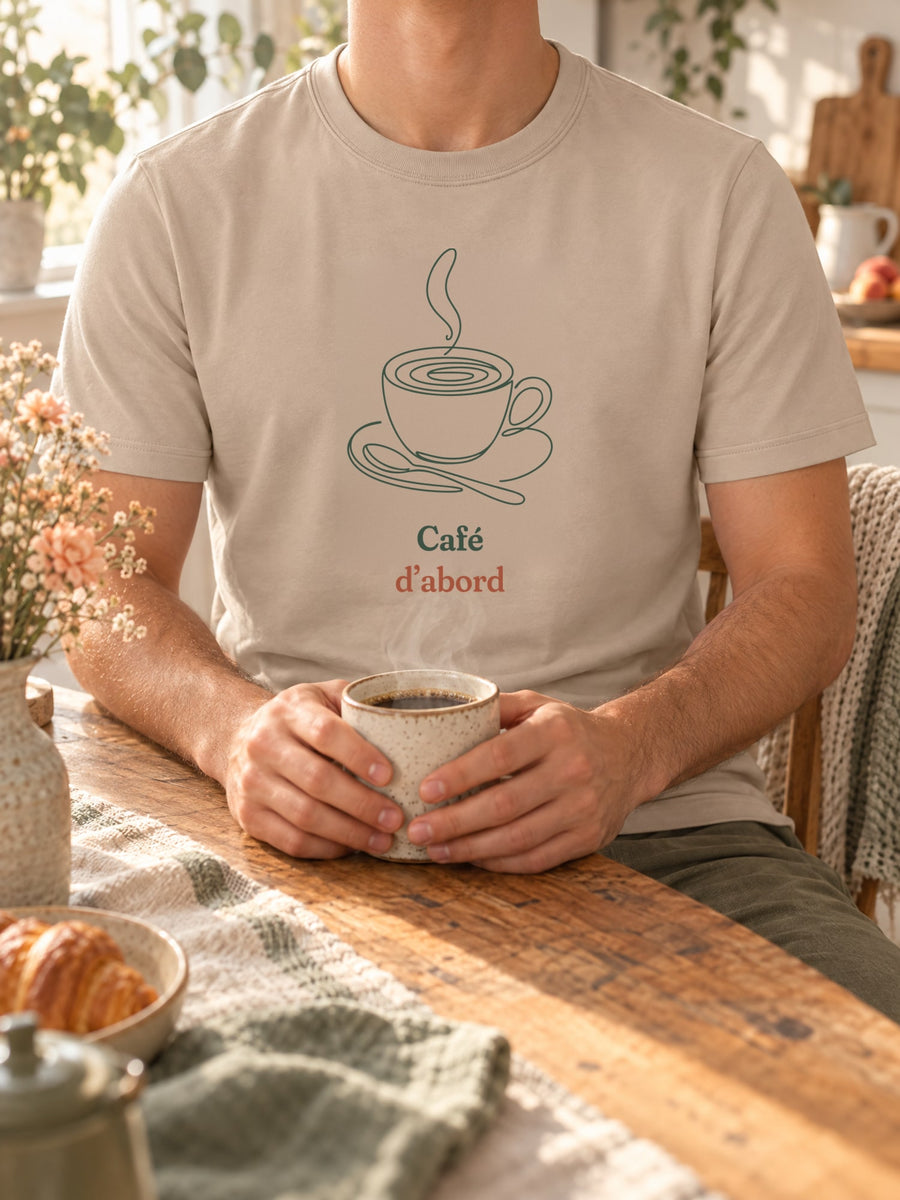 Visuel d’illustration – t-shirt dans une mise en situation, avec le motif une tasse de café fumante sur sa soucoupe, avec sa cuillère, dessinée d’un seul trait continu et « Café d’abord »