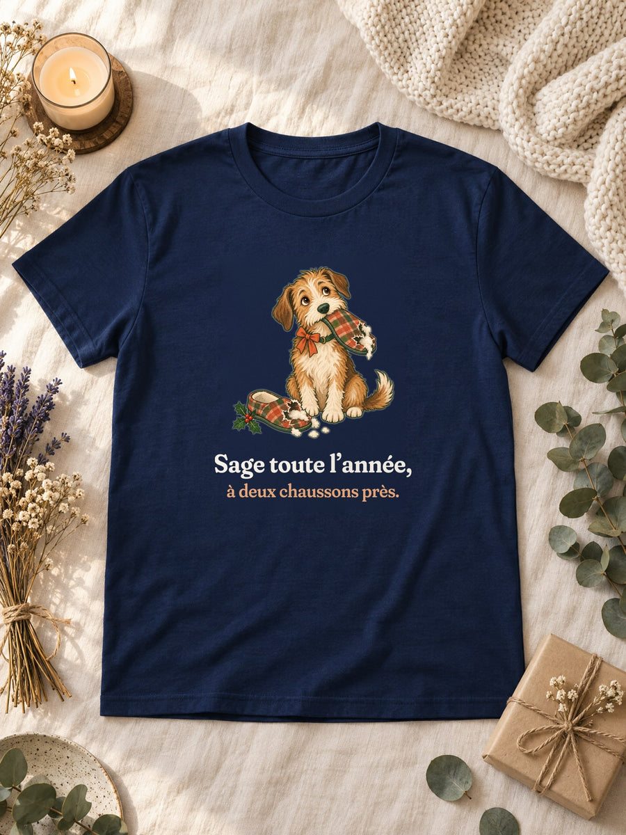 Visuel d’illustration – t-shirt navy posé à plat sur du lin, avec des fleurs séchées, une bougie et un paquet cadeau, avec le motif de Noël : un chien assis, un chausson dans la gueule, et la phrase « Sage toute l’année, à deux chaussons près. »