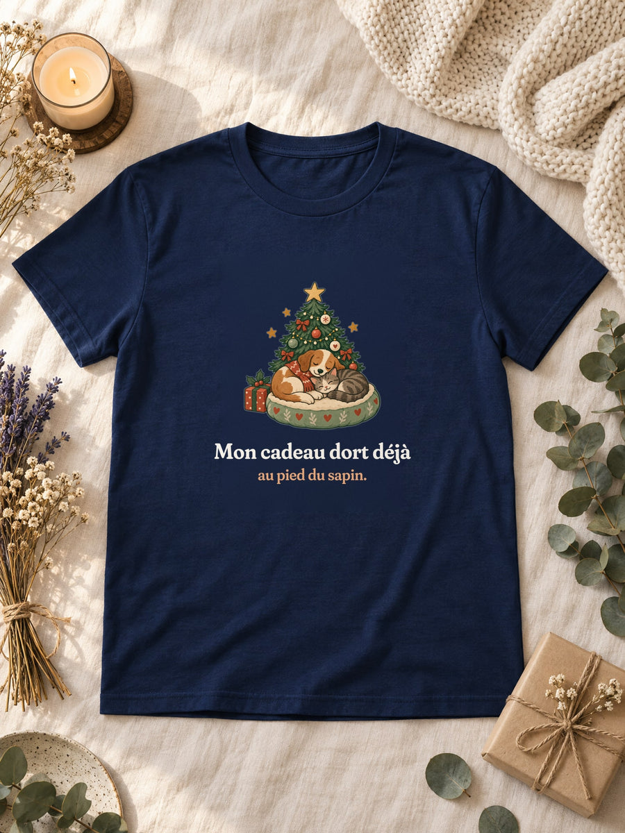 Visuel d’illustration – t-shirt navy posé à plat sur du lin, avec des fleurs séchées, une bougie et un paquet cadeau, avec le motif de Noël : un chien et un chat endormis au pied d’un petit sapin et la phrase « Mon cadeau dort déjà au pied du sapin. »