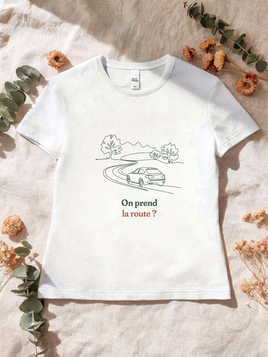 Visuel d’illustration – T-shirt femme blanc avec le motif une voiture sur une route sinueuse entre les arbres, dessinée d’un seul trait continu et « On prend la route ? »