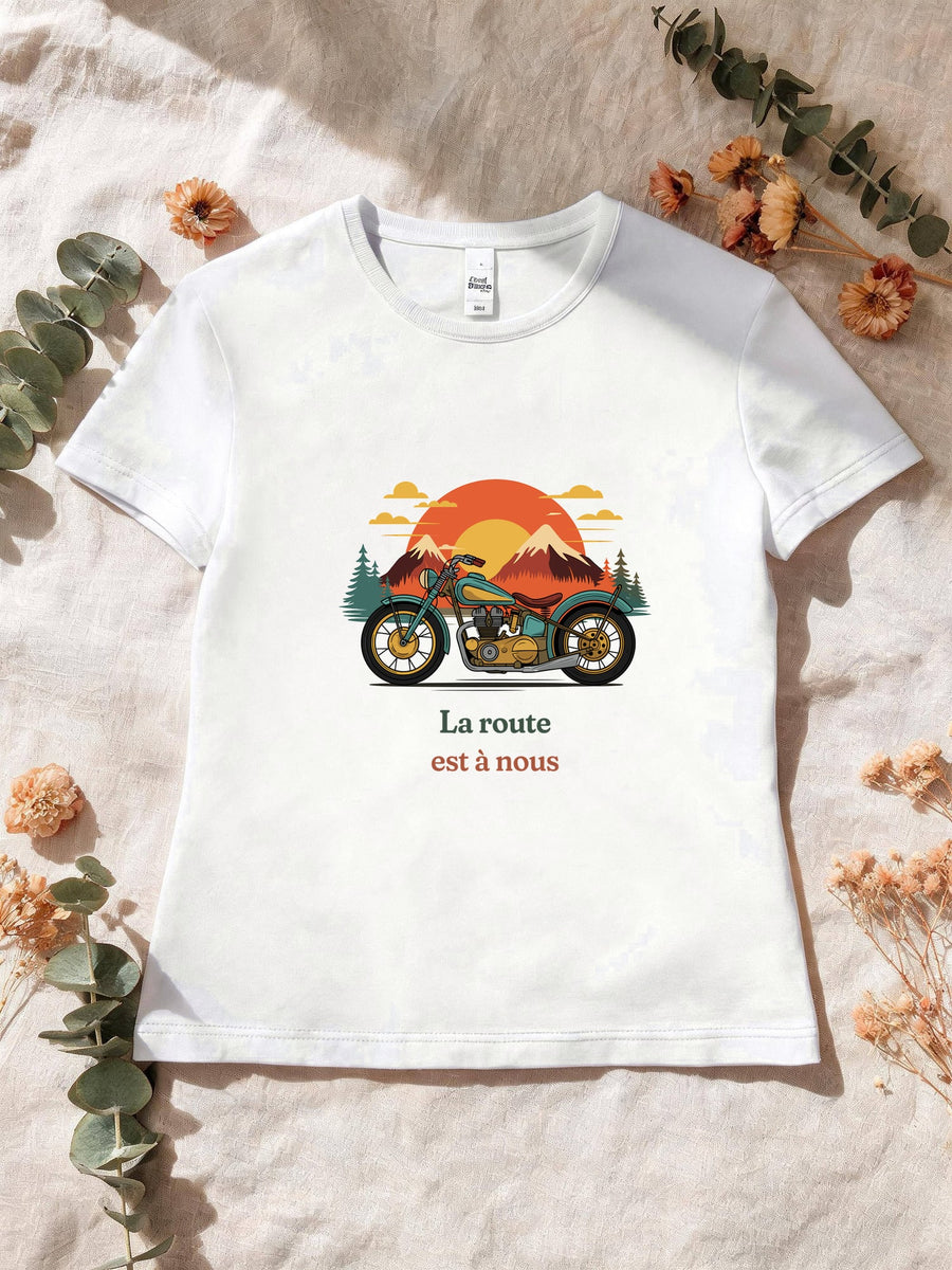 Visuel d’illustration – T-shirt femme blanc avec le motif une moto ancienne illustrée en couleurs devant des montagnes, des sapins et un soleil couchant et « La route est à nous »